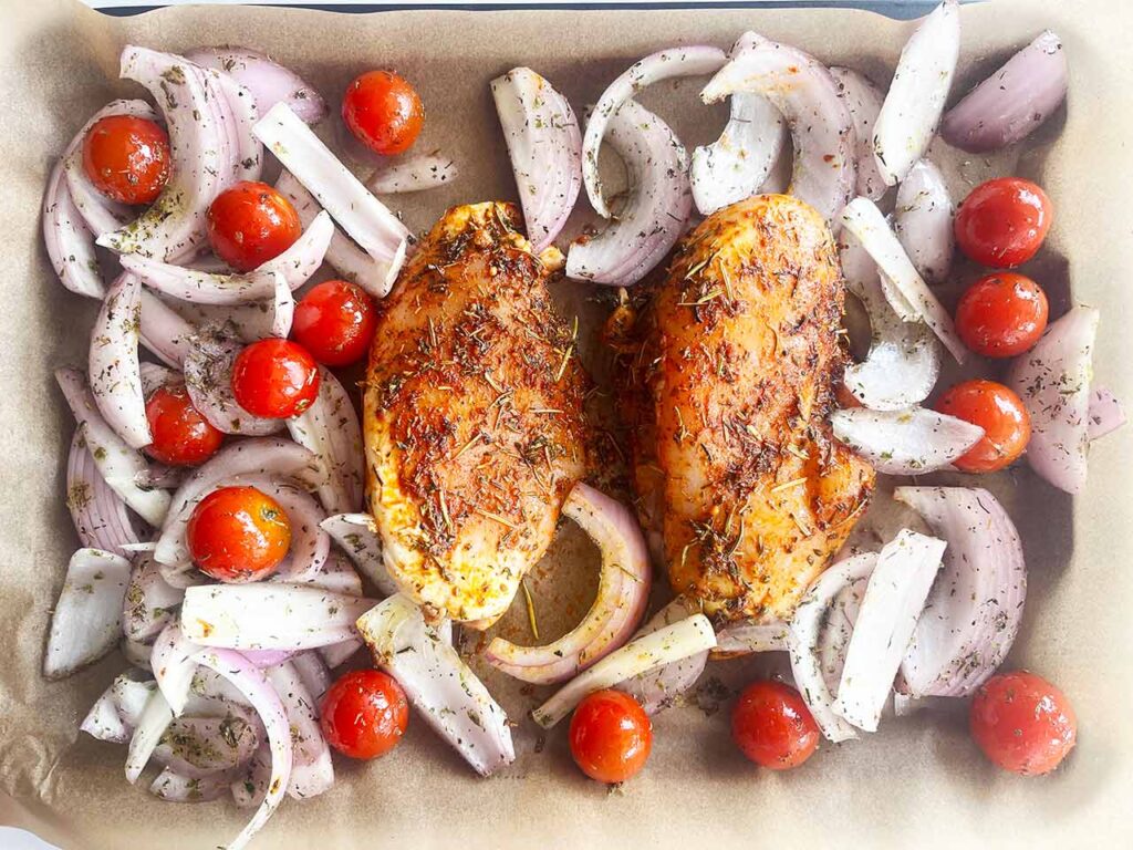 Raw veggies and two raw, seasoned chicken breasts on a parchment-lined sheet pan. Overhead focus.
