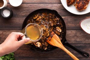 Adding chicken broth to a skillet filled with sautéed mushrooms and onions.