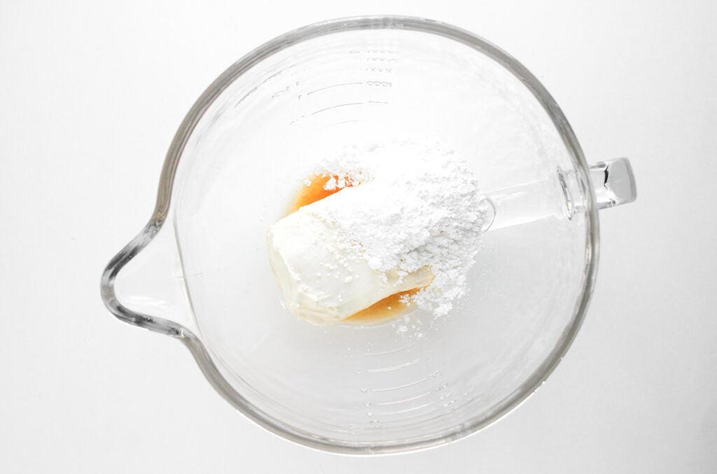 Cream cheese, powdered sweetener, and vanilla extract in a blender tumbler. Overhead focus.