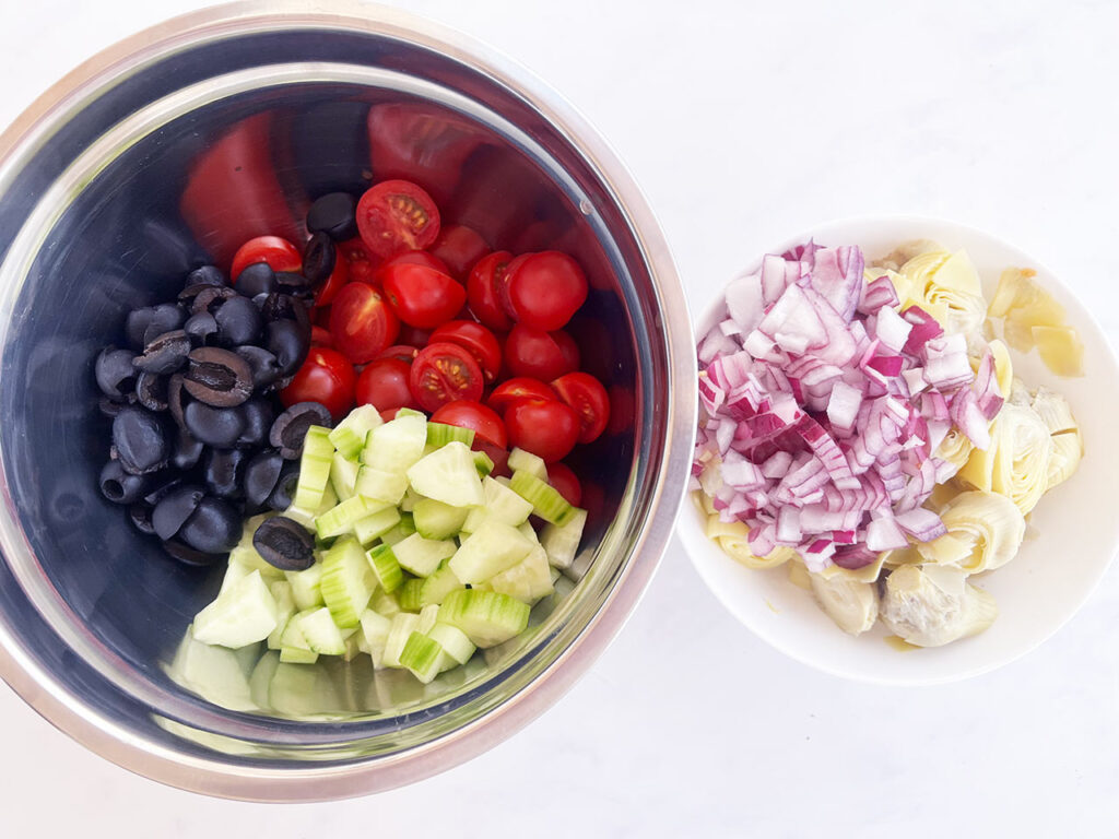 Two bowls. One with olives, tomatoes and cucumber. The other with chopped onions and artichoke hearts. Overhead focus.