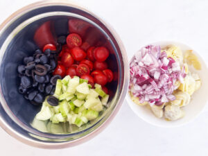 Two bowls. One with olives, tomatoes and cucumber. The other with chopped onions and artichoke hearts. Overhead focus.