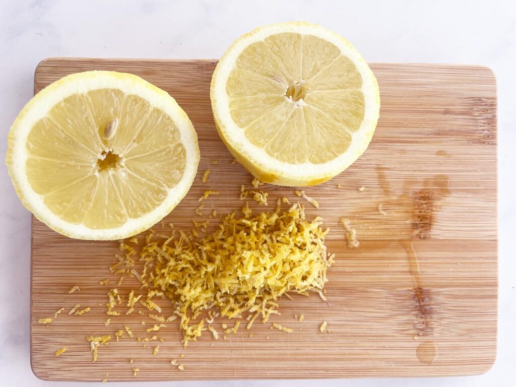 A cut lemon on a wood cutting board next to a pile of lemon zest.