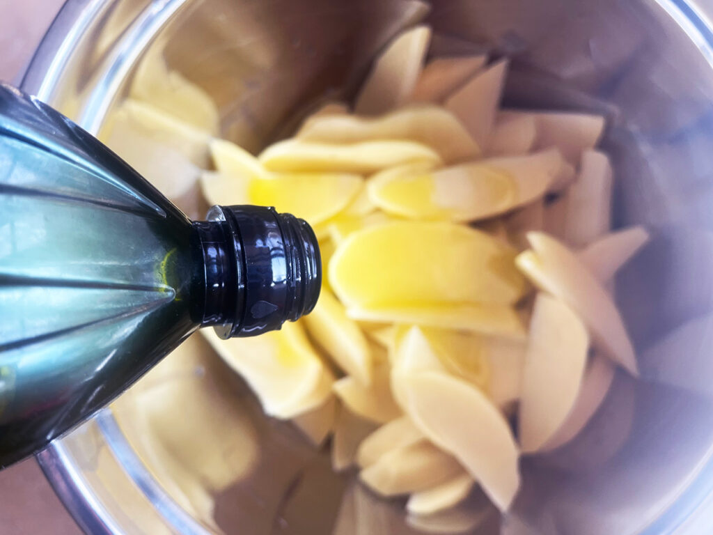 Adding oil to potato wedges in a metal mixing bowl.