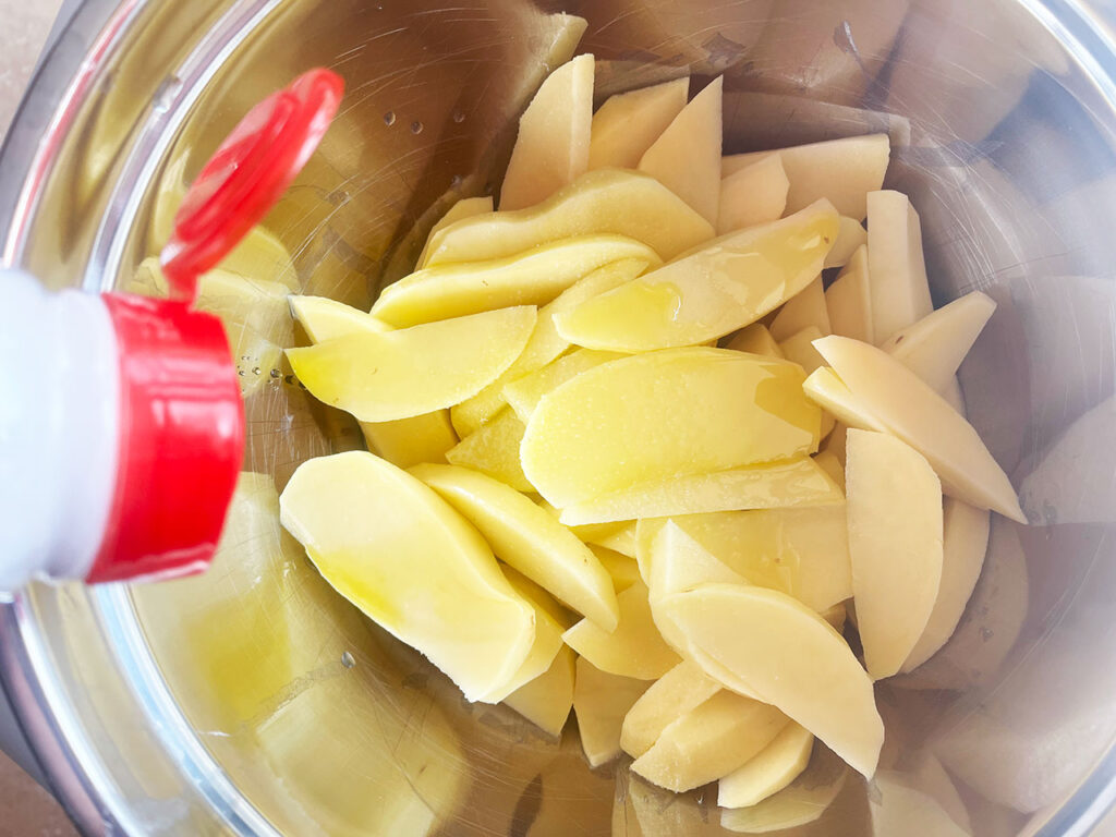 Adding salt to potato wedges and oil in a metal mixing bowl.
