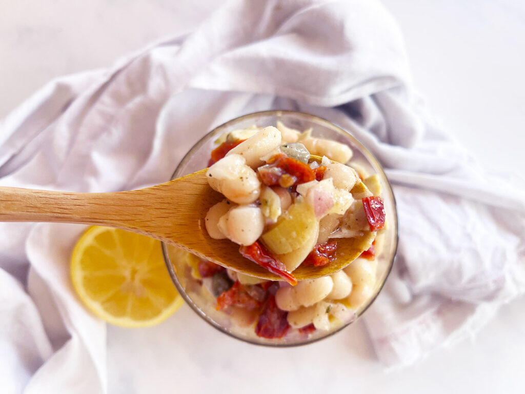 Overhead view. A wood spoon lifts up a bite of Tuscan White Bean Salad from a jar filled with it.