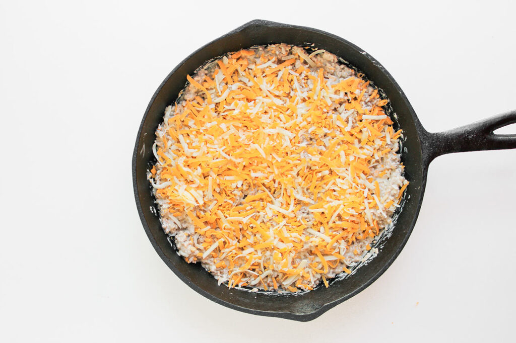 Shredded cheese sprinkled over the top of ground meat and cream cheese mix in a cast iron skillet.