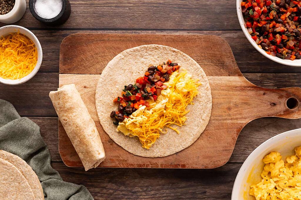 A tortilla on a wood cutting board with cheese, eggs, and vegetable mix lined up on it.