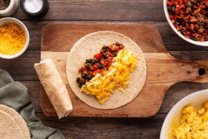 A tortilla on a wood cutting board with cheese, eggs, and vegetable mix lined up on it.