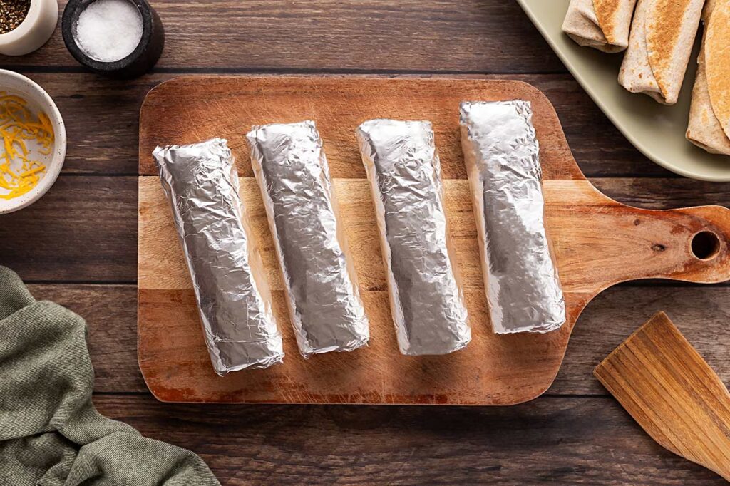 Four Healthy Breakfast Burritos wrapped in foil, laying on a wood cutting board.
