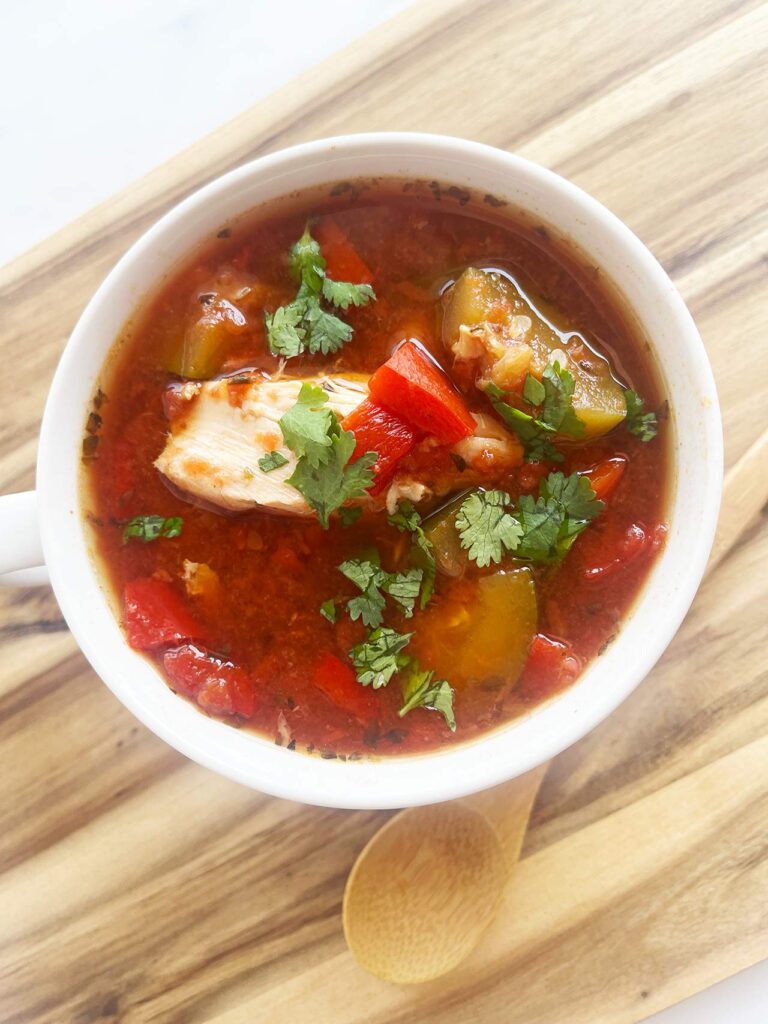 Overhead focus. A white mug filled with Mediterranean Chicken Soup sitting on a wood surface.