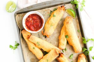 Overhead view of Chicken Taquitos on a baking pan with red dipping sauce in a small, white bowl.