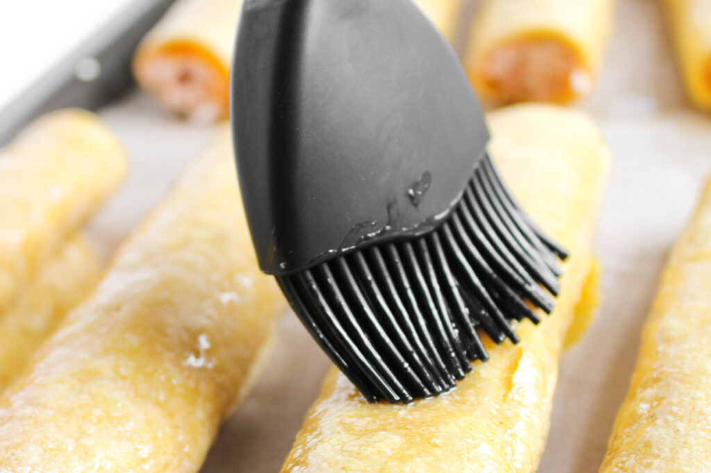 Brushing oil over the taquitos on the baking pan before baking.
