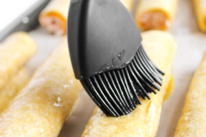 Brushing oil over the taquitos on the baking pan before baking.