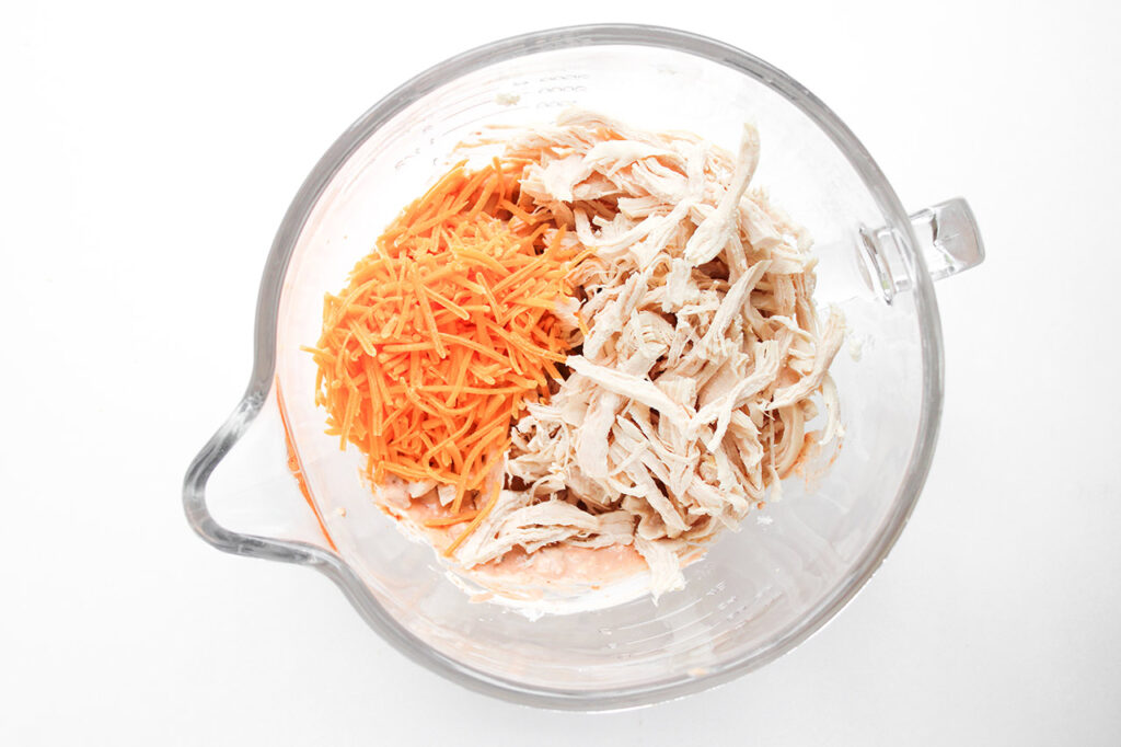 Shredded chicken and shredded cheddar added to a mixing bowl with cream cheese and seasoning.