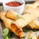 Front view of chicken taquitos on a baking pan. A bowl or red salsa sits at the back of the pan.