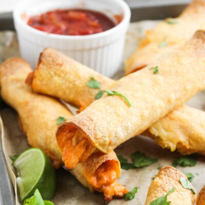Front view of chicken taquitos on a baking pan. A bowl or red salsa sits at the back of the pan.