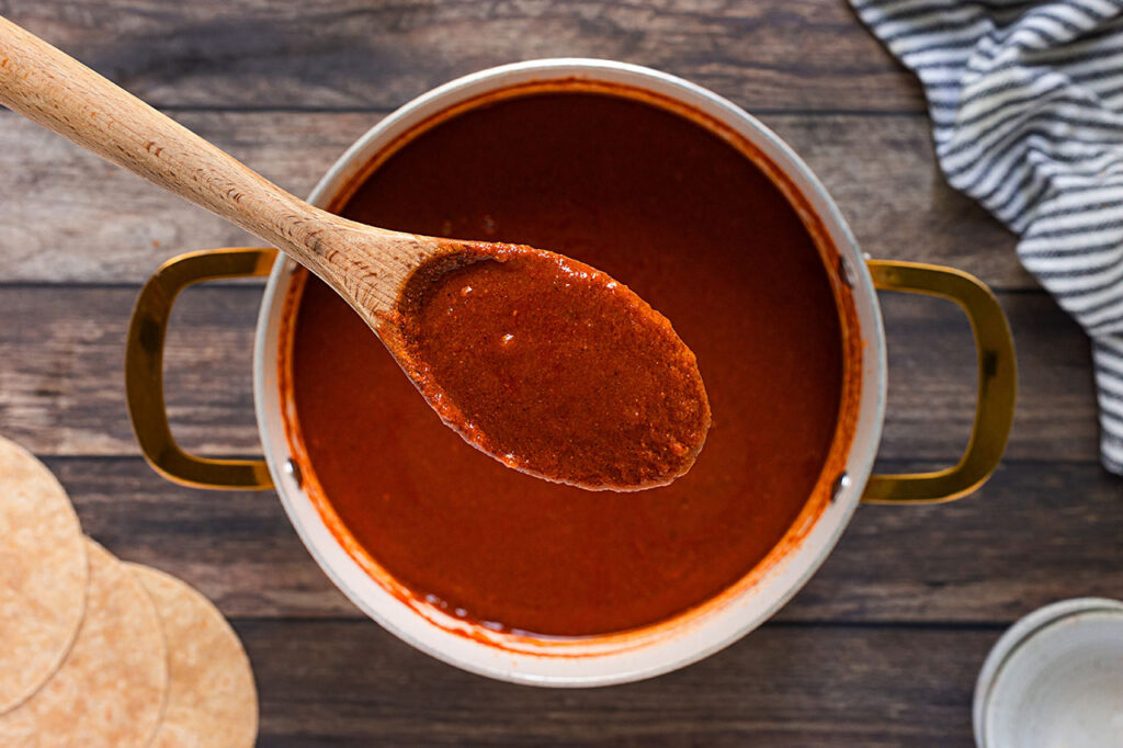 A wood spoon lifts some Enchilada Sauce from a pot full of it. Overhead focus.
