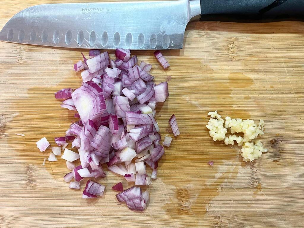 Chopped red onion and minced garlic on a cutting board with a knife.