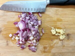 Chopped red onion and minced garlic on a cutting board with a knife.