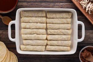 A white casserole dish filled with just-rolled tortillas stuffed with beans and chicken.