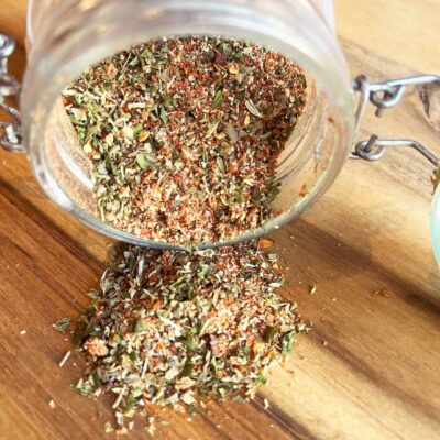 Front view of an open jar of Copycat Trader Joe's Pizza Seasoning, tipped over on a wood cutting board.