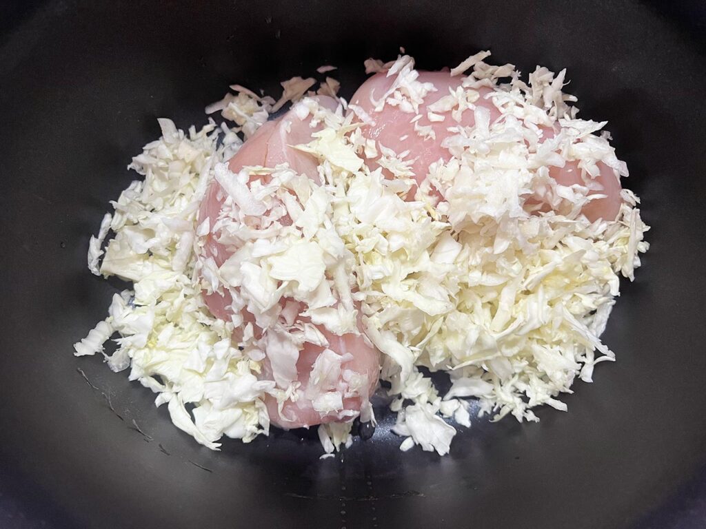 Shredded cabbage added to chicken breasts in a slow cooker crock.