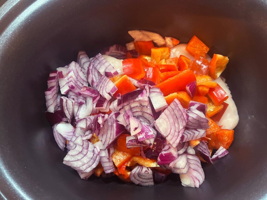 Chopped red onion added to peppers and chicken in a slow cooker crock.