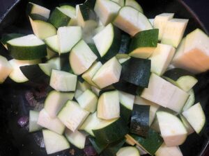 Chopped zucchini added to a skillet with onions and spices.