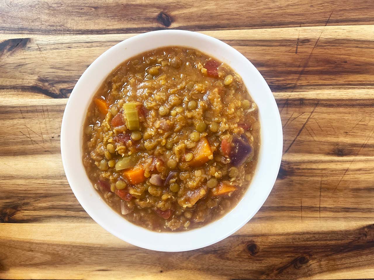Overhead focus. A white bowl on a wood surface, filled with slow cooker lentil stew.