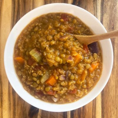 Overhead focus. A small, wood spoon rests in a white bowl of slow cooker lentil stew.