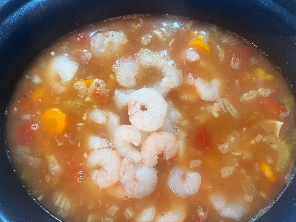 Overhead focus. A slow cooker crock with cooked veggies and broth. Raw shrimp are added.