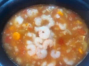 Overhead focus. A slow cooker crock with cooked veggies and broth. Raw shrimp are added.