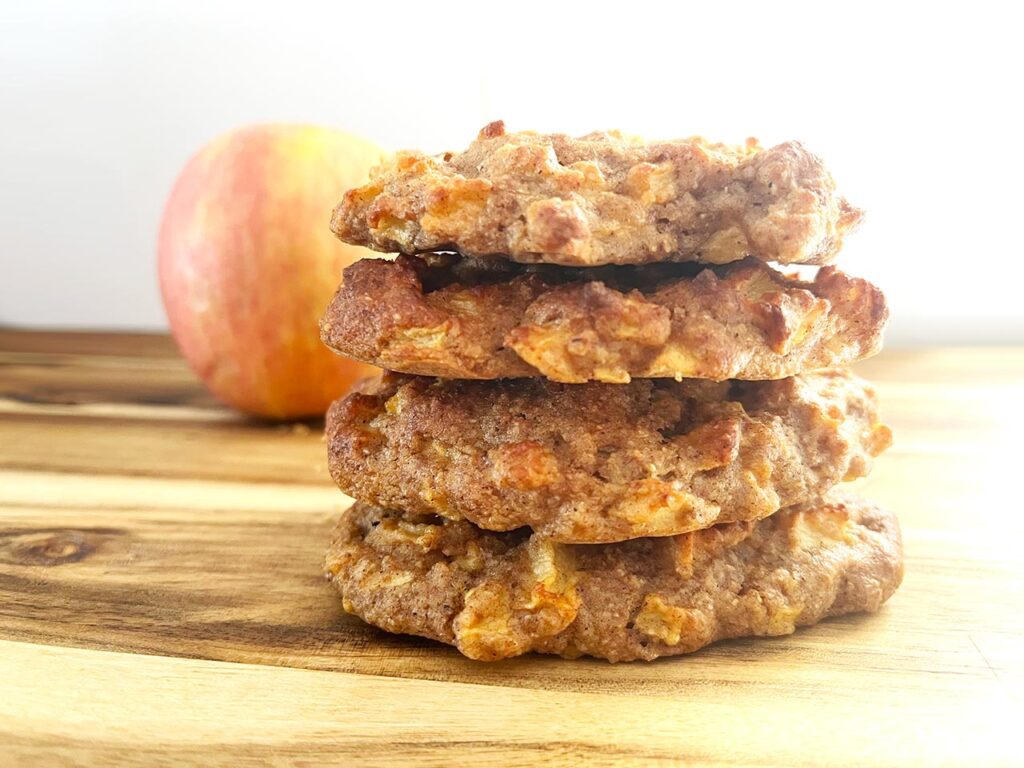 A stack of apple cookies on a wood surface with a whole apple behind it.