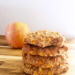 Front focus. A stack of apple cookies sits on a wood surface with an apple behind it and to the left.