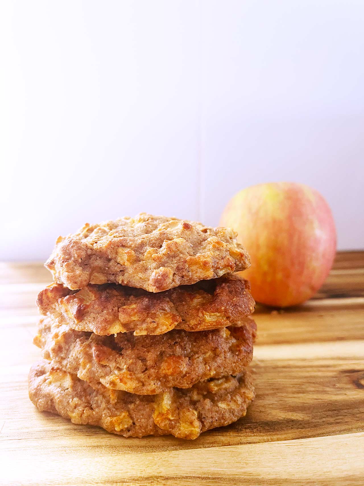 Front focus. A stack of four apple cookies on a wood surface. An apple sits behind them to the right.