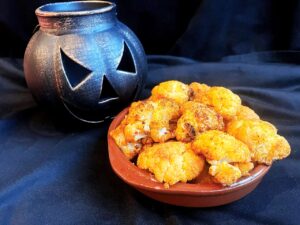 A small plate of roasted cauliflower brains sits on a black surface with a metal jack-o-lantern sitting behind it.
