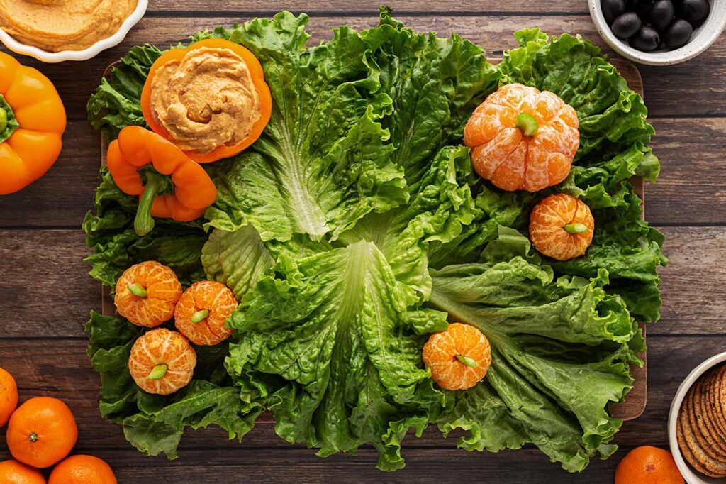 A charcuterie board covered in flat, green lettuce leaves. Mandarin pumpkins and a bell pepper with hummus fill the edges.