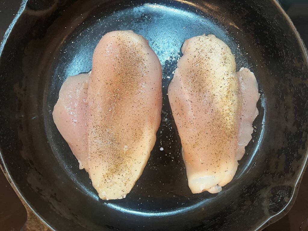Two chicken breasts seasoned with salt and pepper, in a skillet.