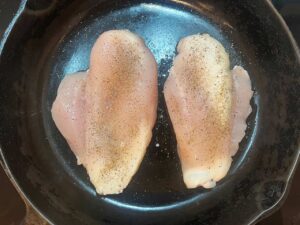 Two chicken breasts seasoned with salt and pepper, in a skillet.