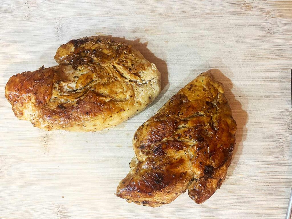 Two cooked chicken breasts on a wood cutting board.