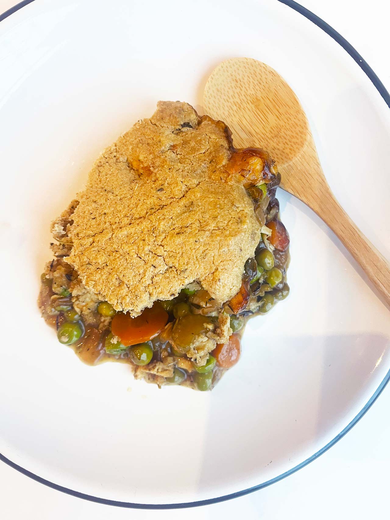 Overhead focus. A serving of chicken cobbler on a white plate. A wood spoon lays next to the food.