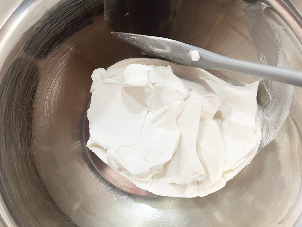 A package of cream cheese in a metal mixing bowl with a spatula.