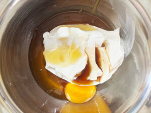 Egg, honey, and vanilla extract added to cream cheese in a metal mixing bowl.