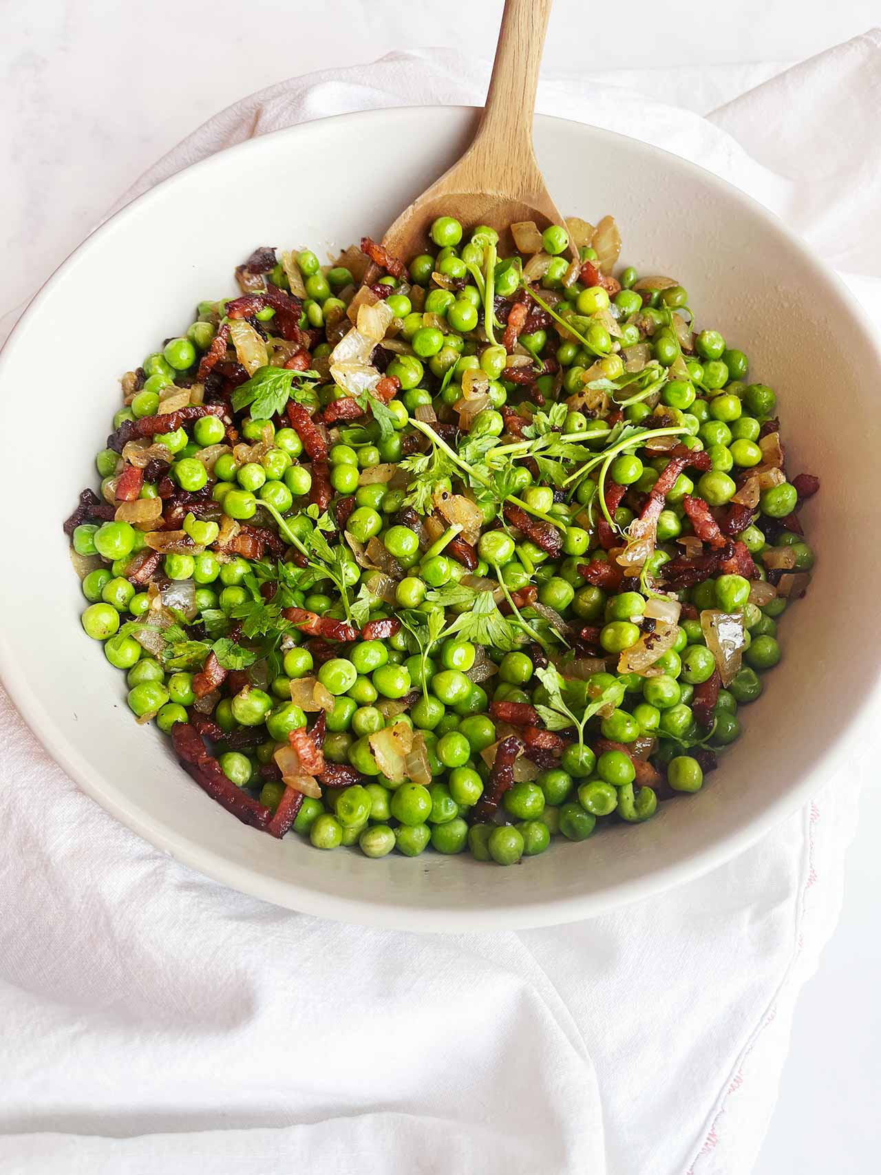 Angled overhead focus. A white bowl with a wood spoon filled with pea salad.