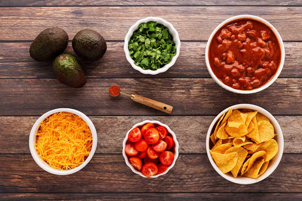 Chili nacho ingredients in individual bowls on a wood surface.