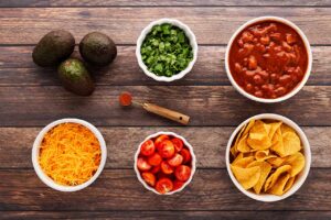 Chili nacho ingredients in individual bowls on a wood surface.