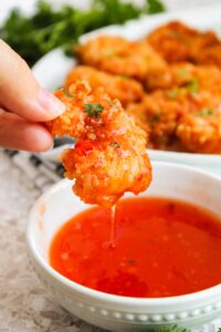A hand holds a Crispy Coconut Shrimp just dipped in chili sauce