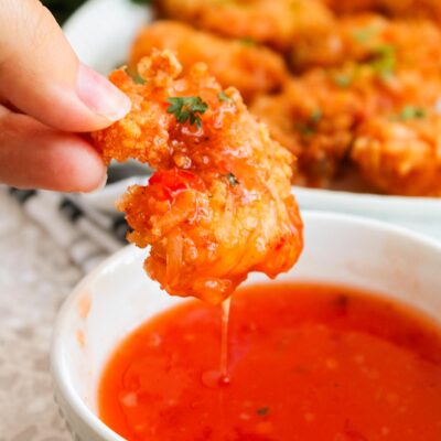 A hand holds a Crispy Coconut Shrimp just dipped in chili sauce