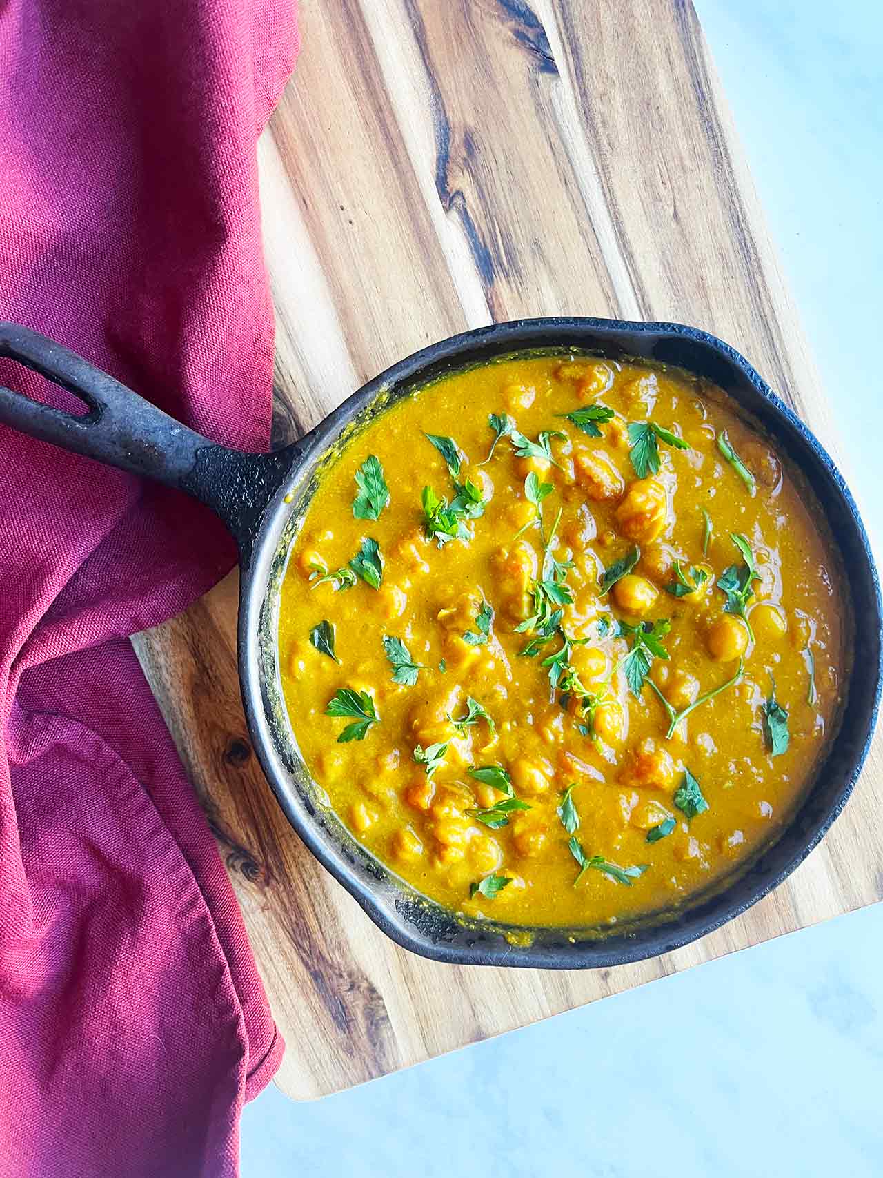 Overhead focus. A black skillet filled with Pumpkin Curry sitting on a wood cutting board with a burgundy-colored towel.