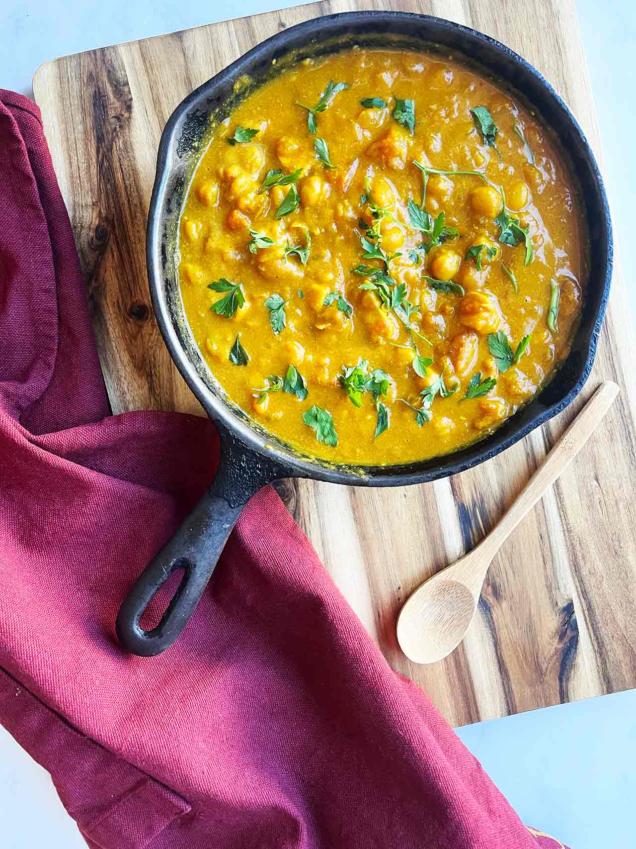 Pumpkin Curry in a black skillet sitting on a wood cutting board with a towel and wood spoon.